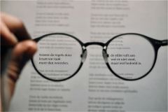 Een persoon houdt een paar ronde, zwarte brillen tegen een blad met kleine, uitgelijnde tekst; de letters door de glazen zijn scherp leesbaar.