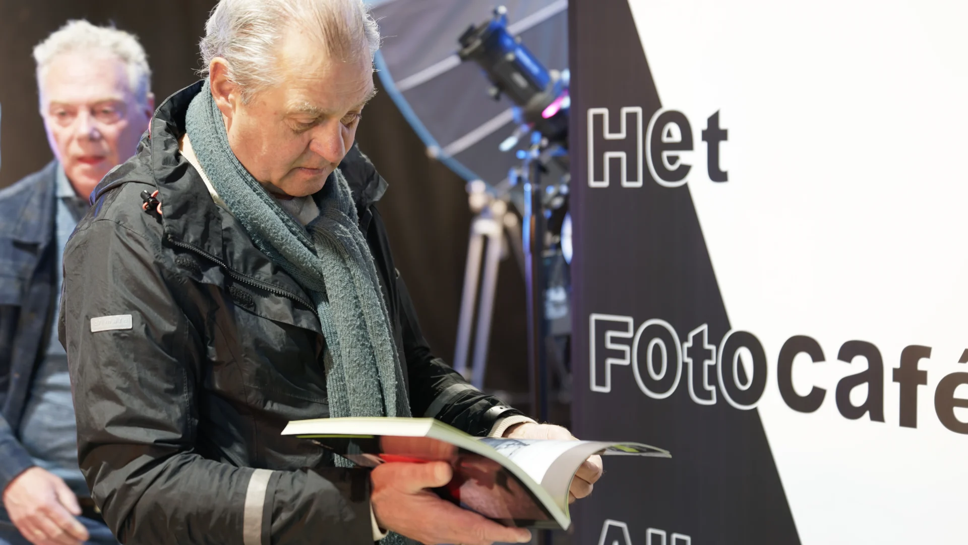 Twee oudere mannen bij een affiche Het Fotocafé Alkmaar, één bekijkt een boek.